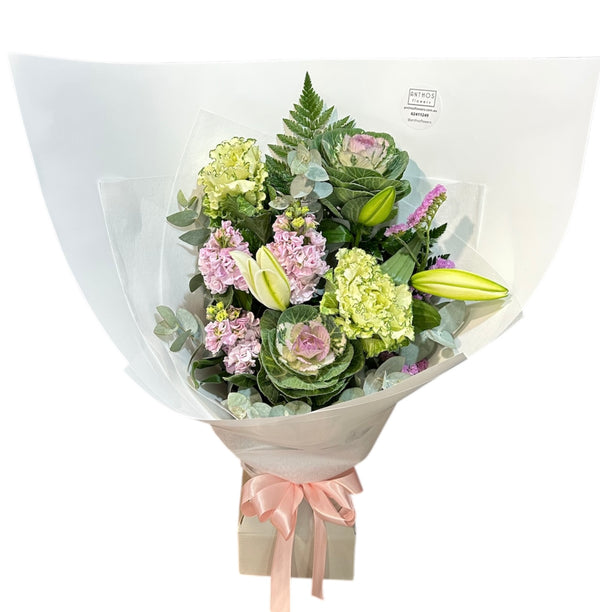 A floral bouquet featuring a variety of flowers including Oriental Lily, Ornamental Kale and Stocks, wrapped in a pastel paper with a pink ribbon.